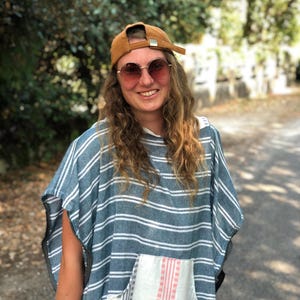 Op de afbeelding: Een bruine honkbalpet met een zilveren gesp, een ronde getinte zonnebril en een blauw-witte gestreepte poncho met een witte voorzak met verticale rode strepen. Het kledingstuk heeft een los, mouwloos ontwerp met een wijd, gedrapeerd silhouet.