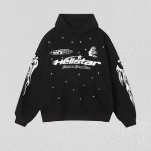 May include: A black long-sleeve hooded sweatshirt featuring white graphic prints. The front displays the text Hellstar with a star icon, a smaller oval logo, and scattered white dots. Both sleeves feature white flame-like graphic designs.