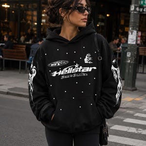 May include: A black hooded sweatshirt featuring white graphic prints and scattered white dots. The front displays the text Hellstar and Faith to Salvation with flame and star motifs. The sleeves feature white flame designs, and the garment includes a front kangaroo pocket.