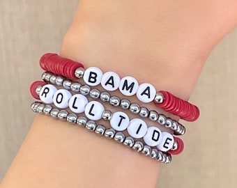May include: Four stacked bracelets with white letter beads spelling "BAMA" and "ROLL TIDE". The bracelets feature red disc beads and silver metallic beads. The bracelets are worn on a wrist.