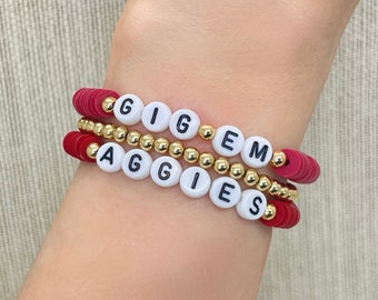 May include: Three stacked bracelets with red disc beads, gold beads, and white letter beads spelling "GIGEM" and "AGGIES". The bracelets are on a wrist, showcasing the jewelry's design and color scheme.
