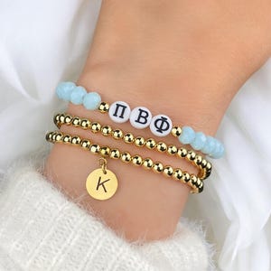 May include: Three bracelets stacked on a wrist. The top bracelet has light blue beads and white letter beads spelling out Greek letters. The middle and bottom bracelets are gold beaded. The bottom bracelet has a gold charm with the letter "K".