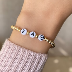 May include: A gold beaded bracelet with white beads that spell out "KAΘ".
