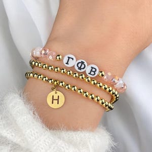 May include: Three beaded bracelets stacked on a wrist. One bracelet features gold beads and a gold charm with the letter "H". Another has white letter beads spelling out Greek letters. The top bracelet has pink crystal beads.