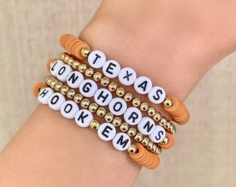 May include: Stack of five handmade bracelets. Each bracelet features white letter beads spelling "TEXAS", "LONGHORNS", and "HOOK EM". The bracelets have gold beads and orange disc beads. The bracelets are stacked on a wrist.
