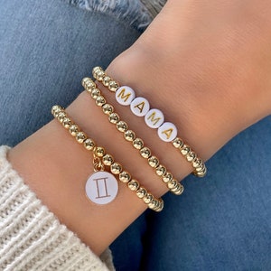 May include: Three gold beaded bracelets. One bracelet spells "MAMA" in white letter beads. Another bracelet has a Gemini charm. The bracelets are worn on a wrist with a white sweater and blue jeans.
