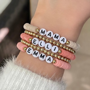 May include: A stack of four bracelets with gold beads and white letter beads spelling out the names "EMMA", "ELLI", and "MAMA". The bracelets are on a person's wrist.