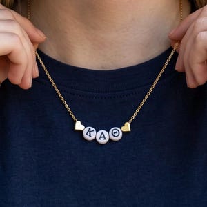 May include: A gold-toned chain necklace featuring three white circular beads with black lettering spelling KAO, flanked by two small gold-toned heart-shaped charms. The necklace rests against a dark navy blue fabric background.