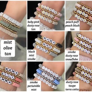 May include: A set of five bracelets with gold beads and white letter beads spelling out names. The bracelets are stacked on a wrist and are in various colors, including tan, olive, black, smoke, dusty rose, periwinkle, and terra cotta.