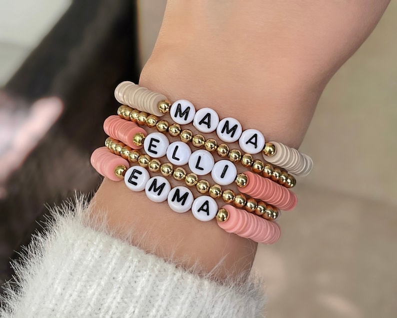 May include: A stack of four bracelets with gold beads and white letter beads spelling out the names "EMMA", "ELLI", and "MAMA". The bracelets are on a person's wrist.