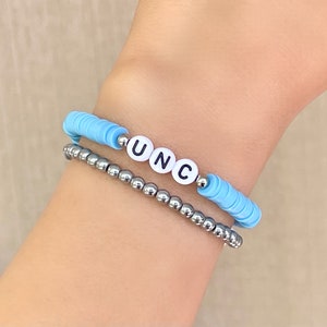 Puede incluir: Dos pulseras apiladas en una mu&ntilde;eca. La pulsera superior es de color azul claro con cuentas de letras blancas que deletrean "UNC". La pulsera inferior est&aacute; hecha de peque&ntilde;as cuentas de color plateado.