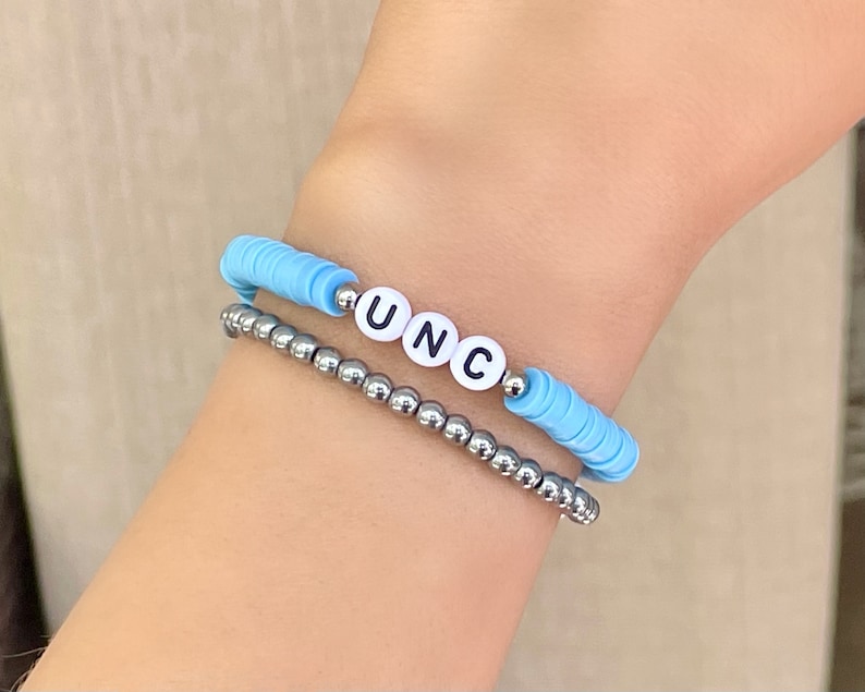 Puede incluir: Dos pulseras apiladas en una mu&ntilde;eca. La pulsera superior es de color azul claro con cuentas de letras blancas que deletrean "UNC". La pulsera inferior est&aacute; hecha de peque&ntilde;as cuentas de color plateado.