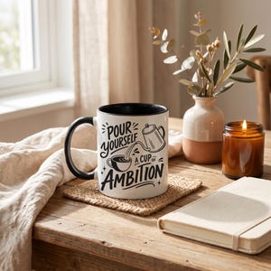 Puede incluir: Una taza de cer&aacute;mica blanca con interior y asa negros, que presenta un texto gr&aacute;fico negro que dice "POUR YOURSELF A CUP OF AMBITION" junto a una ilustraci&oacute;n de una cafetera vertiendo l&iacute;quido en una taza, decorada con peque&ntilde;os detalles de estrellas.
