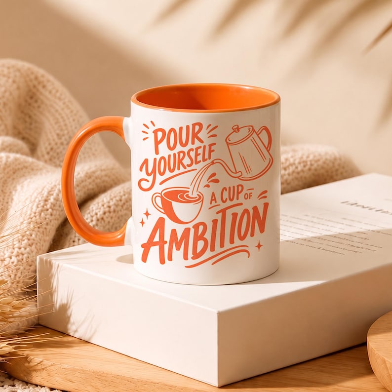 Puede incluir: Una taza de cer&aacute;mica blanca con interior y asa de color naranja. El exterior presenta un texto naranja que dice "POUR YOURSELF A CUP OF AMBITION" junto a una ilustraci&oacute;n gr&aacute;fica de una cafetera vertiendo l&iacute;quido en una taza peque&ntilde;a.