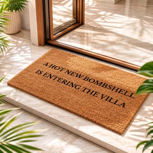 May include: A rectangular tan coir doormat featuring the black text A HOT NEW BOMBSHELL IS ENTERING THE VILLA printed in a bold, sans-serif font across the center. The mat has a coarse, fibrous texture and is positioned on a light-colored floor.