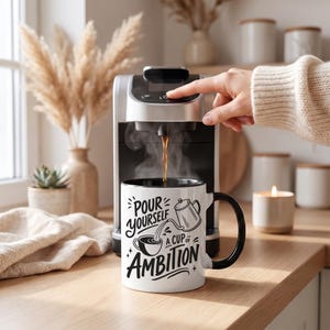 Puede incluir: Una taza de cer&aacute;mica blanca con asa y borde negros, con el texto en negro Pour Yourself A Cup Of Ambition y un gr&aacute;fico de una cafetera y una taza, colocada bajo una cafetera plateada y negra que est&aacute; sirviendo caf&eacute; caliente.