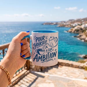 Puede incluir: Una taza de cer&aacute;mica blanca con interior y asa de color azul brillante. El exterior presenta un texto en azul oscuro que dice "POUR YOURSELF A CUP OF AMBITION" junto a una ilustraci&oacute;n gr&aacute;fica de una cafetera vertiendo l&iacute;quido.