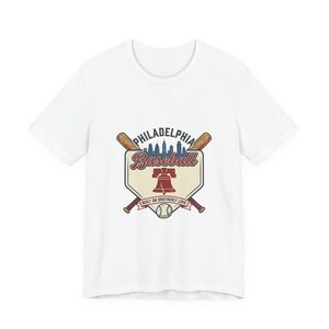 Puede incluir: Una camiseta blanca de manga corta con un gráfico de una base de béisbol, dos bates de madera cruzados, una pelota de béisbol y la Liberty Bell. El texto dice Philadelphia, Baseball y Built on Brotherly Love, con la silueta de un horizonte urbano.