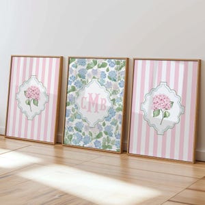 May include: Three framed wall art prints. The two outer prints feature pink and white vertical stripes with a central white quatrefoil emblem containing a pink hydrangea. The center print displays a blue floral pattern with a central monogram CMB.