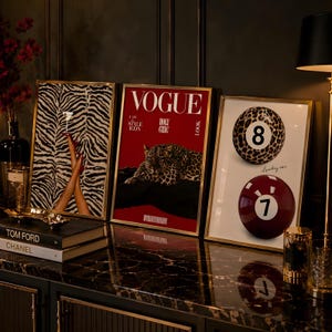 May include: Three framed wall art prints in gold frames. The left print shows legs in red heels against a zebra pattern. The centre print is a red Vogue magazine cover with a leopard. The right print features a leopard print 8-ball and a red 7-ball.