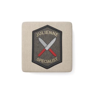 Peut inclure: Un dessous de verre carré beige avec un écusson hexagonal centré. L'écusson présente un fond texturé gris foncé avec une broderie de deux couteaux argentés croisés aux manches rouges et le texte JULIENNE SPECIALIST en lettres crème.