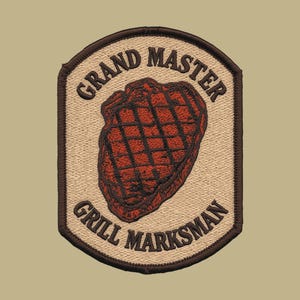 Puede incluir: Un parche bordado en forma de escudo con fondo beige y borde marrón oscuro. En el centro, un filete a la parrilla de color rojo parduzco con marcas de parrilla negras. El texto GRAND MASTER aparece arqueado arriba y GRILL MARKSMAN abajo.