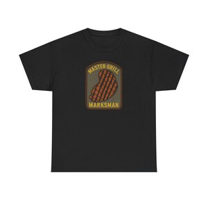 Puede incluir: Una camiseta negra de manga corta con un gráfico rectangular en el pecho. El gráfico muestra un filete a la parrilla de color marrón con marcas de cocción, rodeado por un borde oscuro y el texto amarillo "MASTER GRILL" arriba y "MARKSMAN" abajo.