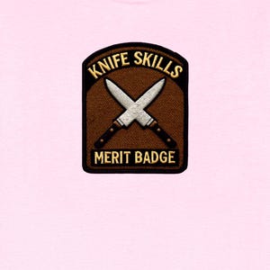 May include: A light pink t-shirt featuring a rectangular embroidered patch with a brown background and a black border. The patch displays two crossed silver knives with dark handles and the text KNIFE SKILLS MERIT BADGE in gold lettering.