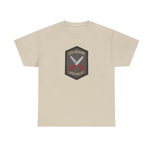 May include: A beige short-sleeve t-shirt featuring a centered dark gray hexagonal patch. The patch displays two crossed knives with red handles and silver blades, with the text JULIENNE above and SPECIALIST below in light-colored lettering.