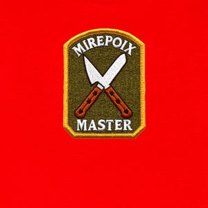 May include: A bright red t-shirt featuring a rectangular embroidered patch with an olive green background. The patch displays two crossed silver chef knives with brown handles and the white text MIREPOIX above and MASTER below the knives.