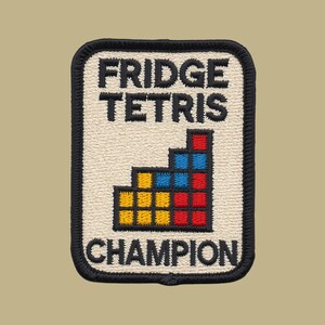 May include: A rectangular embroidered patch with a cream-colored background and a black border. The text reads FRIDGE TETRIS CHAMPION in black block letters. Below the text is a graphic of stacked yellow, red, and blue square blocks.