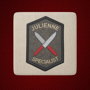 May include: A rectangular off-white coaster featuring a centered hexagonal embroidered patch. The patch has a dark gray background with two crossed silver knives with red handles. The text JULIENNE is above and SPECIALIST is below the knives.