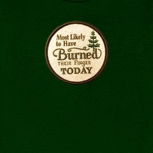 May include: A dark green t-shirt featuring a circular embroidered patch with a cream-colored background and a dark brown border. The patch displays the text "Most Likely to Have Burned THEIR FINGER TODAY" in brown, with a small green pine tree icon.
