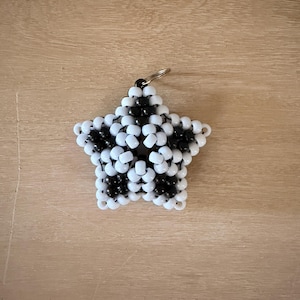 May include: A star-shaped keychain crafted from small, glossy pony beads in alternating black and white colors. The beads are arranged in a geometric pattern to form a five-pointed star, with a silver-toned metal split ring attached at the top.