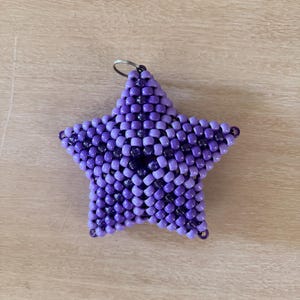 May include: A star-shaped charm crafted from small, interlocking purple and violet beads. The design features a repeating geometric pattern with a mix of opaque light purple and translucent dark violet beads, finished with a small metal ring at the top.
