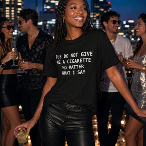 Op de afbeelding: Een zwart T-shirt met korte mouwen en witte tekst die luidt: PLS DO NOT GIVE ME A CIGARETTE NO MATTER WHAT I SAY. Het shirt heeft een ronde hals en een standaard pasvorm, gemaakt van effen zwarte stof met duidelijke, opvallende letters.