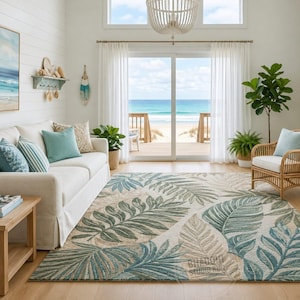 May include: A large rectangular area rug featuring a repeating pattern of oversized tropical palm leaves in shades of teal, sage green, and cream. The rug is positioned on a light wood floor in a living room with a white sofa and a rattan chair.