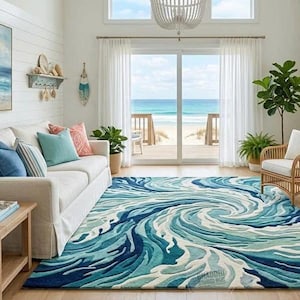 May include: A rectangular area rug featuring a swirling, textured pattern of ocean waves in shades of deep navy, royal blue, turquoise, and white. The rug is positioned on a light wood floor in a room with a white sofa and a view of the ocean.