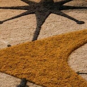 May include: A textured area rug featuring a cream-colored background with a large, dark charcoal starburst shape and a prominent, mustard-yellow boomerang-shaped design. The surface has a thick, tufted pile with visible loops and varied yarn textures.
