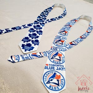 Op de afbeelding: Twee dassen met een witte achtergrond. De ene das heeft een herhalend blauw logo en tekst van de Toronto Maple Leafs. De andere das toont het logo en de tekst van de Toronto Blue Jays in blauw, wit en oranje. Beide dassen hebben een punt.