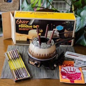 May include: An Oster electric fondue set with a beige and brown pot, a black base with a temperature control knob, and a long handle. The set includes a box, a cookbook, and a tray of metal fondue forks with colored tips, all featuring the Oster brand.