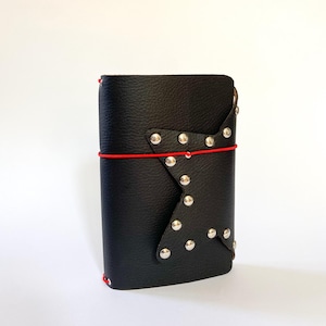 May include: A black leather journal cover with a textured surface, featuring a star-shaped overlay on the front panel adorned with silver-toned metal studs. A thin, vibrant red elastic cord wraps horizontally around the center to secure the cover.