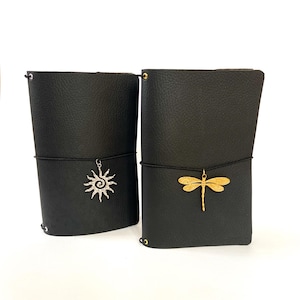 May include: Two black textured journals with elastic closures. The left journal features a silver sun charm, while the right journal features a gold dragonfly charm. Both journals have gold-toned metal eyelets at the top and bottom edges.