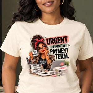 以下が含まれることがあります： クリーム色のTシャツ。デスクで書類の山（Invoices、P.O.s、Approvals、Paymentsと記載）に向かう人物のグラフィックが描かれています。テキストは「URGENT IS NOT A PAYMENT TERM」と書かれています。人物はヒョウ柄のトップスと赤いヘッドバンドを着用しています。