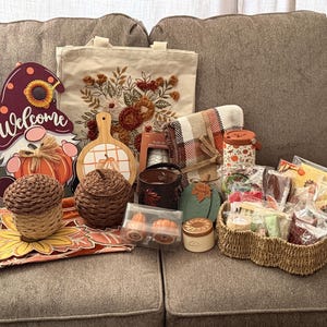 May include: A collection of autumn-themed items including a gnome sign with the word Welcome, a floral embroidered tote bag, a plaid blanket, a woven basket, two acorn-shaped containers, a wooden cutting board, candles, and various small gift items.