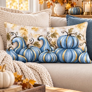 May include: A rectangular throw pillow featuring a printed illustration of multiple blue pumpkins with gold-colored ribs and stems. The background is a cream-colored wash with gold leaf accents and green vine details, creating a seasonal harvest design.