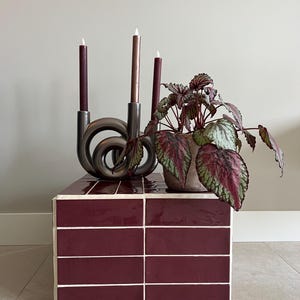 May include: A rectangular side table covered in glossy dark maroon tiles with white grout lines. On top sits a metallic grey sculptural candle holder with three tapered candles in shades of burgundy and copper, alongside a potted begonia plant.