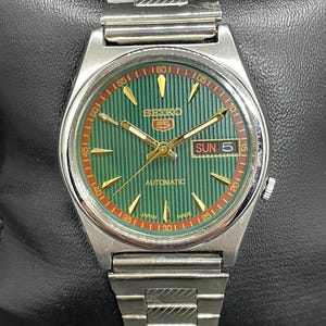 May include: A silver-tone Seiko 5 automatic wristwatch featuring a green dial with vertical pinstripes, gold-tone hands and hour markers, a day-date window at the three o'clock position, and a stainless steel link bracelet with a textured pattern.