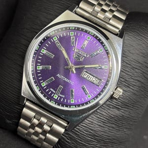 Peut inclure: Une montre-bracelet automatique Seiko 5 argent&eacute;e dot&eacute;e d'un cadran violet &eacute;clatant &agrave; effet rayon de soleil, avec des index rectangulaires, des aiguilles luminescentes et un guichet jour-date &agrave; trois heures. Bracelet &agrave; maillons en acier.