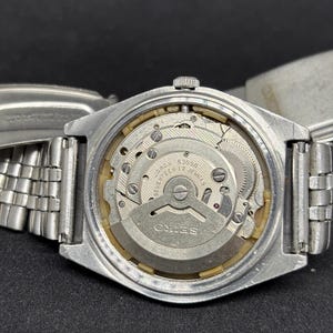 Peut inclure: Une montre-bracelet Seiko argent&eacute;e avec le bo&icirc;tier arri&egrave;re retir&eacute; pour r&eacute;v&eacute;ler le mouvement m&eacute;canique interne. Le m&eacute;canisme en m&eacute;tal porte les inscriptions 'SEIKO', '6309A' et 'SEVENTEEN 17 JEWELS' grav&eacute;es sur la masse oscillante.